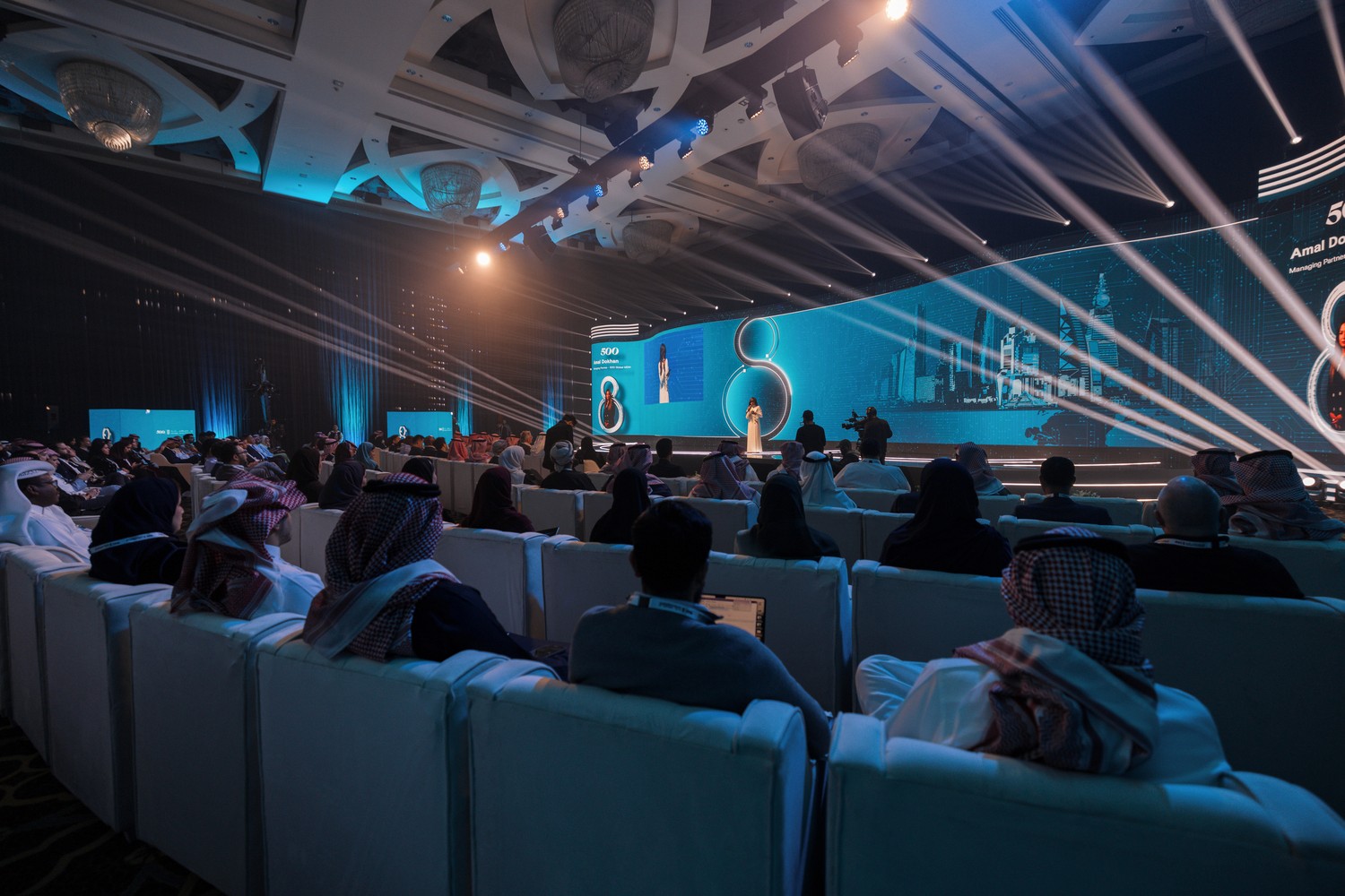جمهور مؤتمر يشاهد متحدثًا على مسرح رقمي داخلي واسع بإضاءة زرقاء