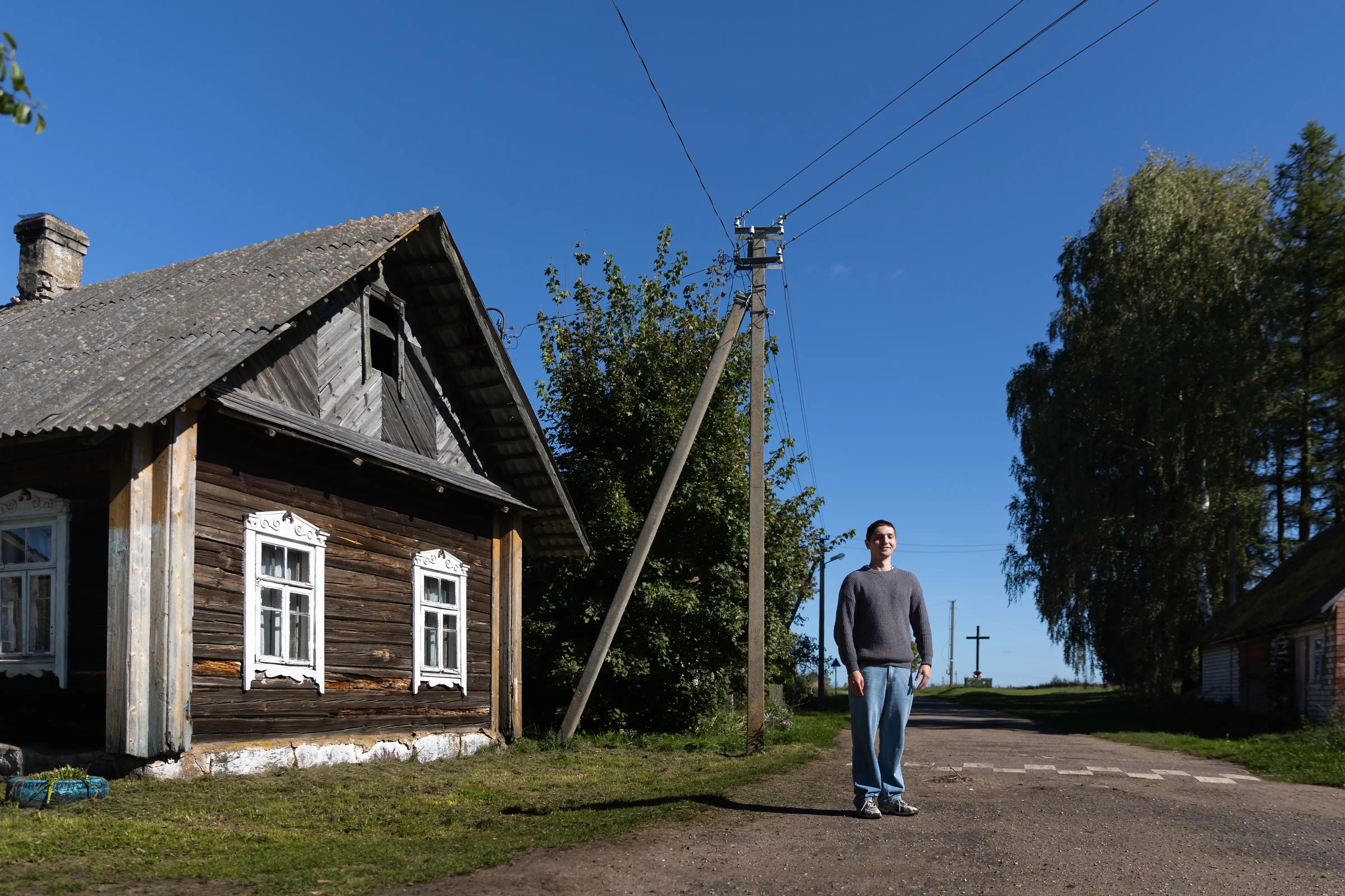 Mężczyzna stoi na drodze obok drewnianego domu w wiejskim otoczeniu. W tle znajduje się niebieskie niebo i drzewa, a na horyzoncie widać krzyż. Linia energetyczna przebiega wzdłuż drogi.