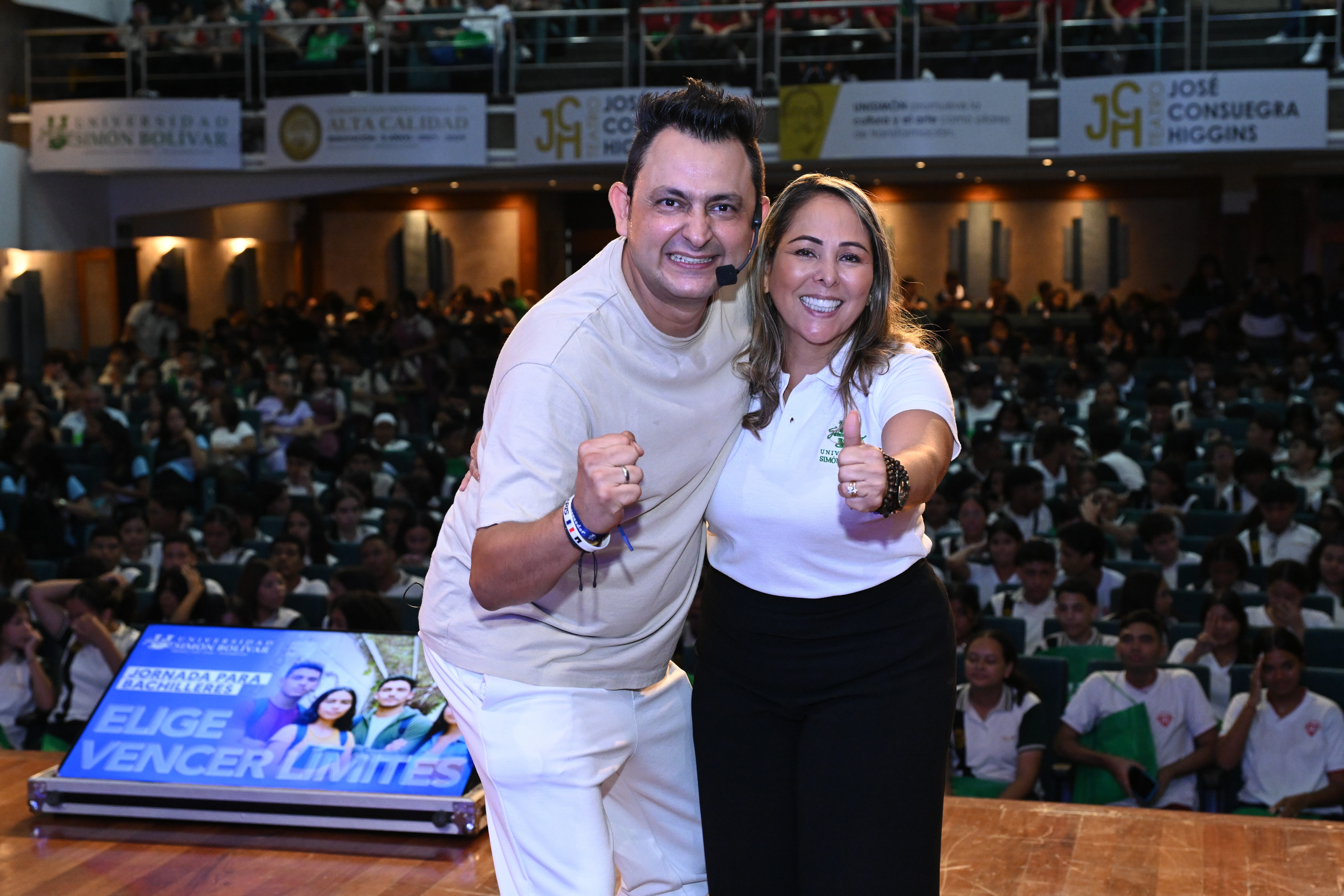 Jorge Baena sonriendo junto a una presentadora en el escenario, ambos haciendo un gesto de fuerza frente a un auditorio lleno de estudiantes durante la conferencia “Elige vencer límites"