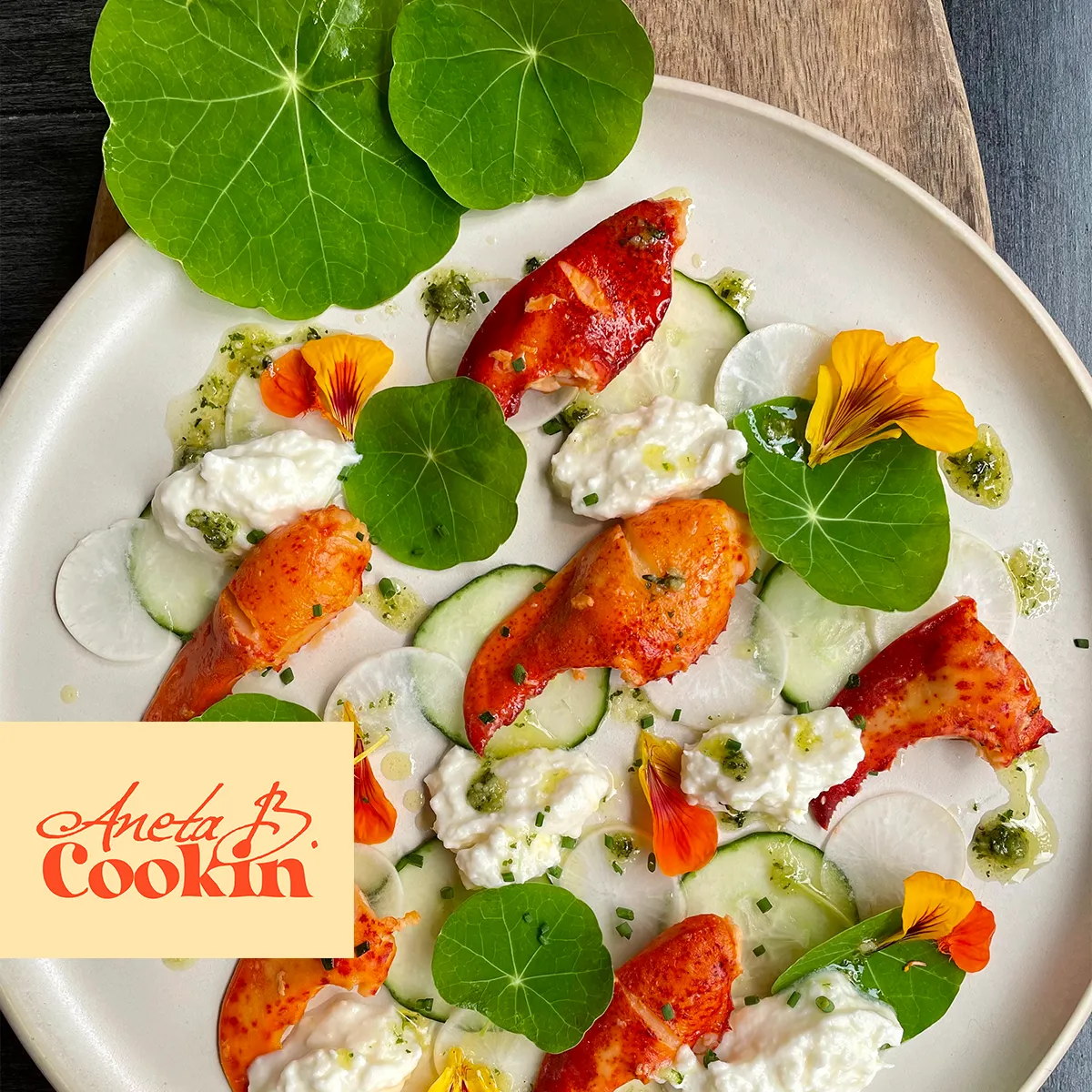 Aneta B. Cookin logo beside an elegant lobster claw dish with edible flowers, showcasing the brand in a fine dining context, designed by The World District.