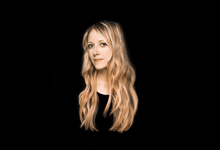 A woman with long, wavy blonde hair is shown against a solid black background. She is looking directly at the camera with a neutral expression. Her hair falls around her shoulders and frames her face. She is wearing a dark-colored top. The lighting is focused on her face and hair.
