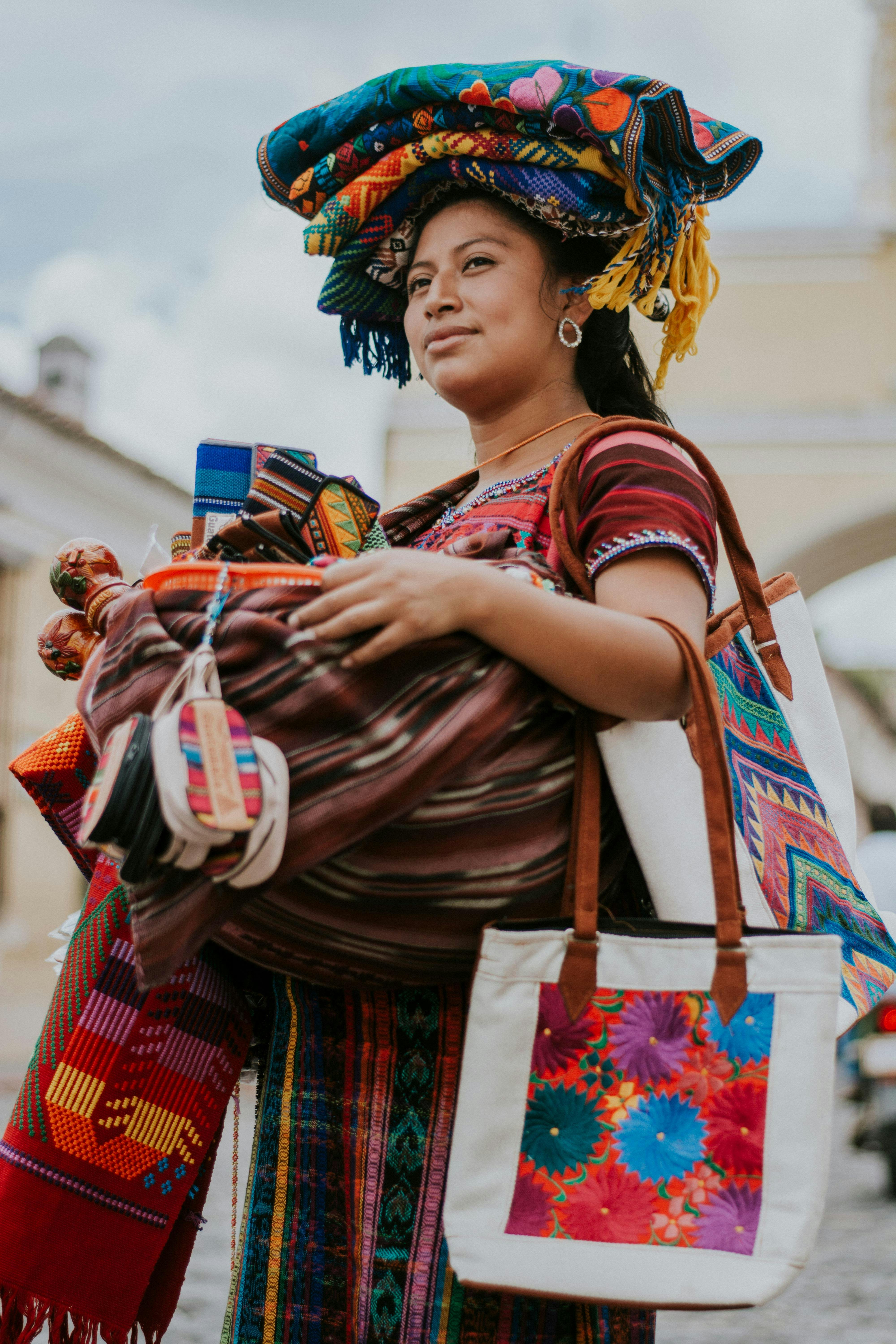A woman in vibrant traditional attire carries a variety of colorful woven textiles and bags, showcasing intricate patterns and rich cultural heritage against a street backdrop.