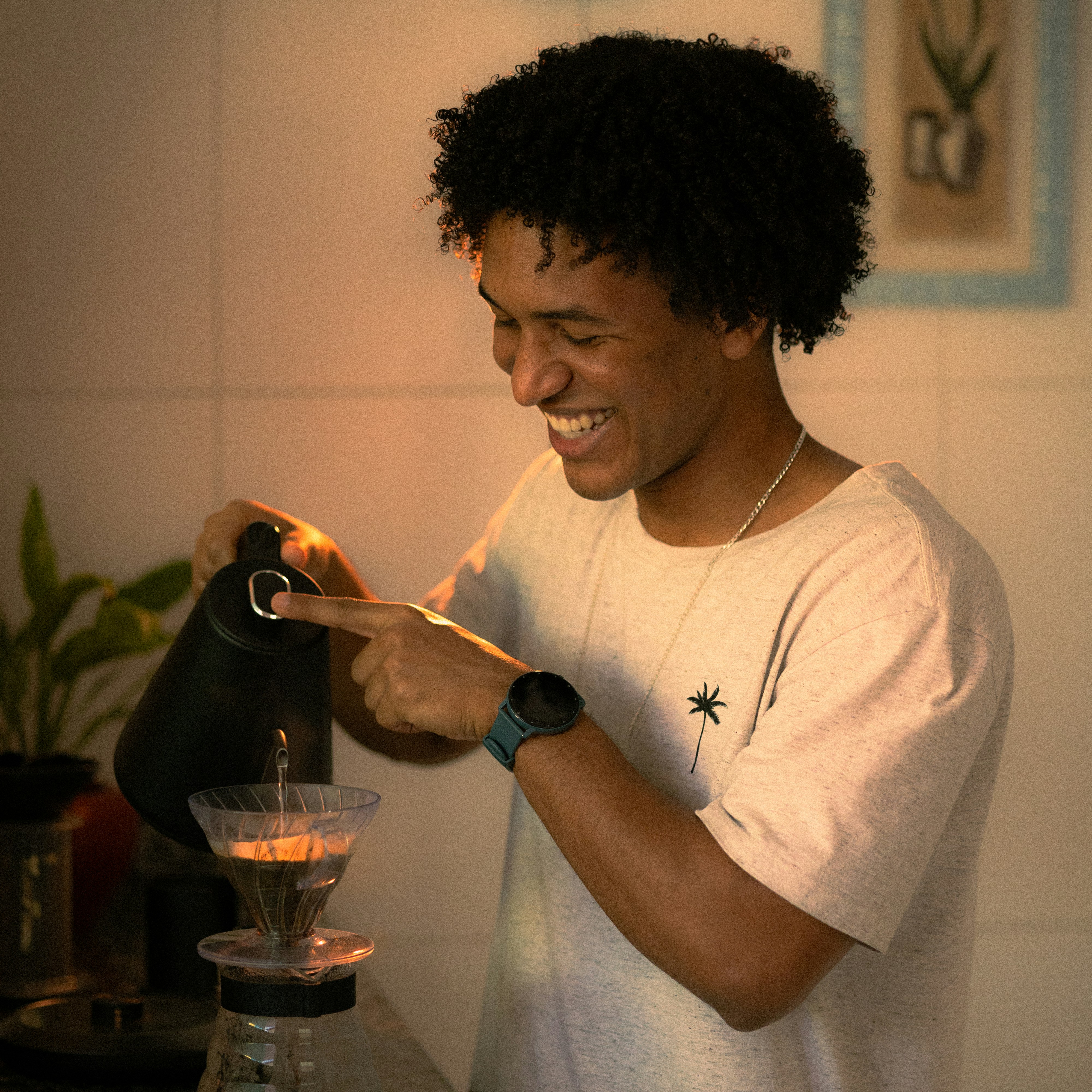 A young person with curly hair smiles at the camera while sitting in front of a laptop against a dark background.