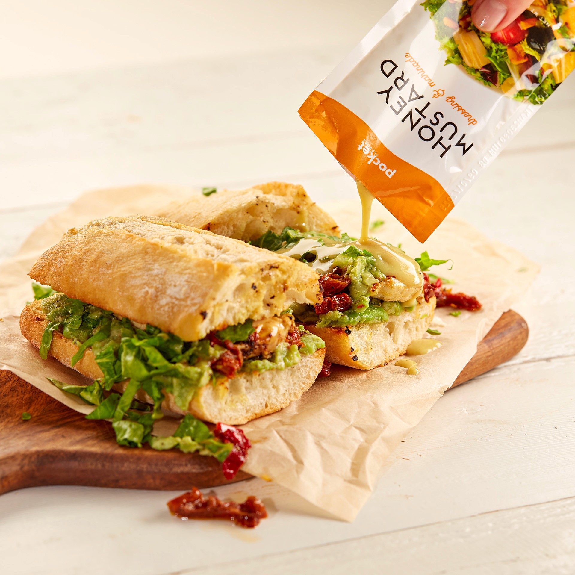 A freshly made sub sandwich on a wooden board is filled with shredded lettuce, sun-dried tomatoes, and slices of chicken, being drizzled with a packet of honey mustard sauce.