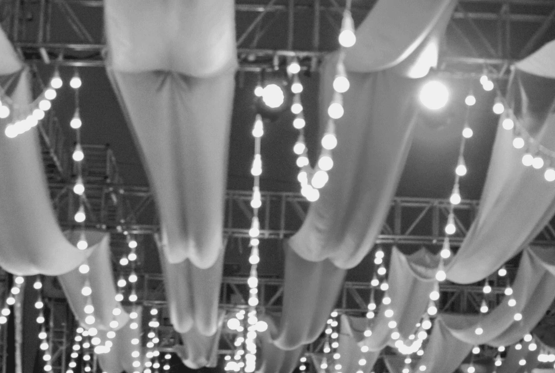 Blurred black-and-white interior of a circus tent lit by soft lantern light; in the center, a subtle graphic outline of a tent is embedded in the composition.