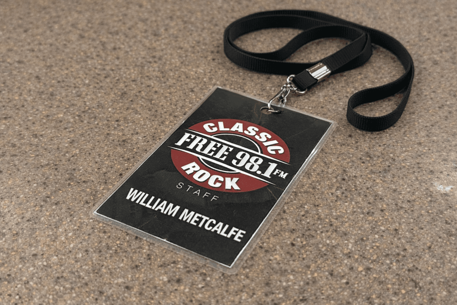 The image shows a staff badge with a lanyard for FREQ 98.1 FM, a Classic Rock radio station. The badge belongs to William Metcalfe, whose name is prominently printed at the bottom. The design features a red, white, and black color scheme, with the words "CLASSIC ROCK" and "STAFF" clearly visible. The badge is placed on a speckled countertop surface.