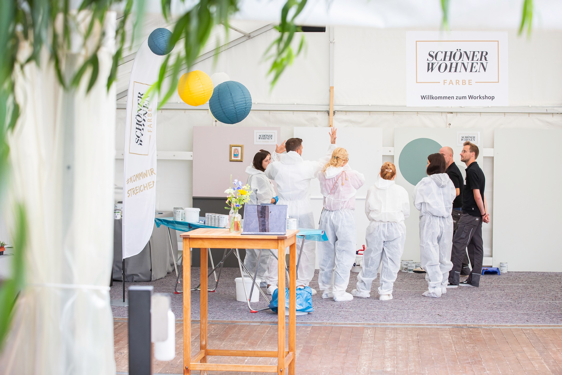 Teilnehmer:innen tragen Schutzanzüge und stehen vor einer Workshop-Fläche mit Farbmuster von Schöner Wohnen Farbe.