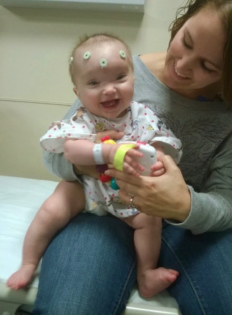 Alt text: Smiling baby with EEG sensors on their head sits on a caregiver’s lap in a medical exam room, holding a toy while the caregiver looks on affectionately.