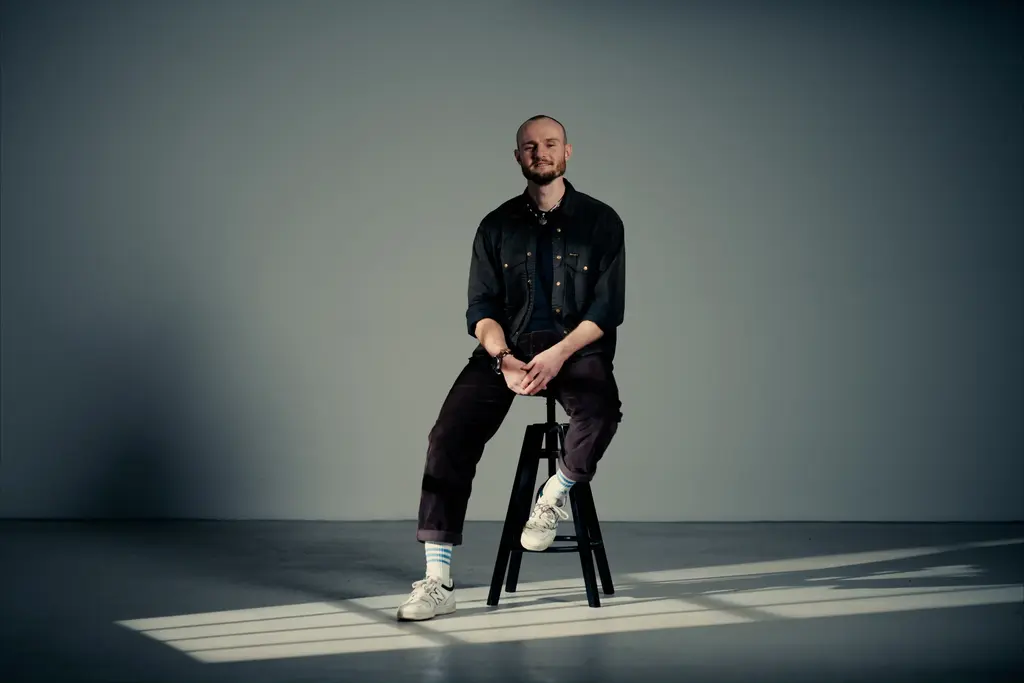 Ein professionelles Porträt von Fabian, einem glücklichen Kunden der Agentur Base and Beyond. Fabian sitzt zufrieden auf einem Hocker in einem professionellen Fotostudio-Setting mit dramatischem Licht und Fensterschatten auf dem Boden. 