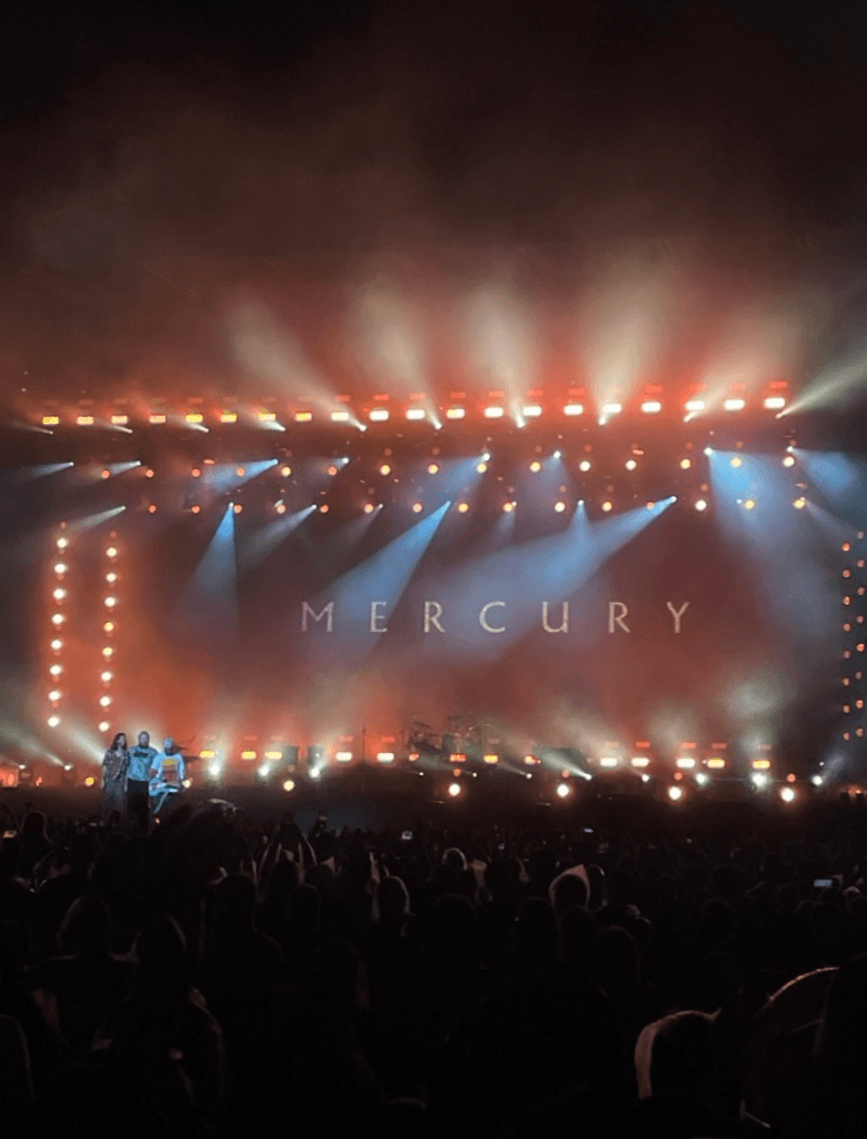 Palco de um show do Image Dragons com iluminação intensa e colorida, com a palavra “MERCURY” exibida no fundo e público olhando para a apresentação.