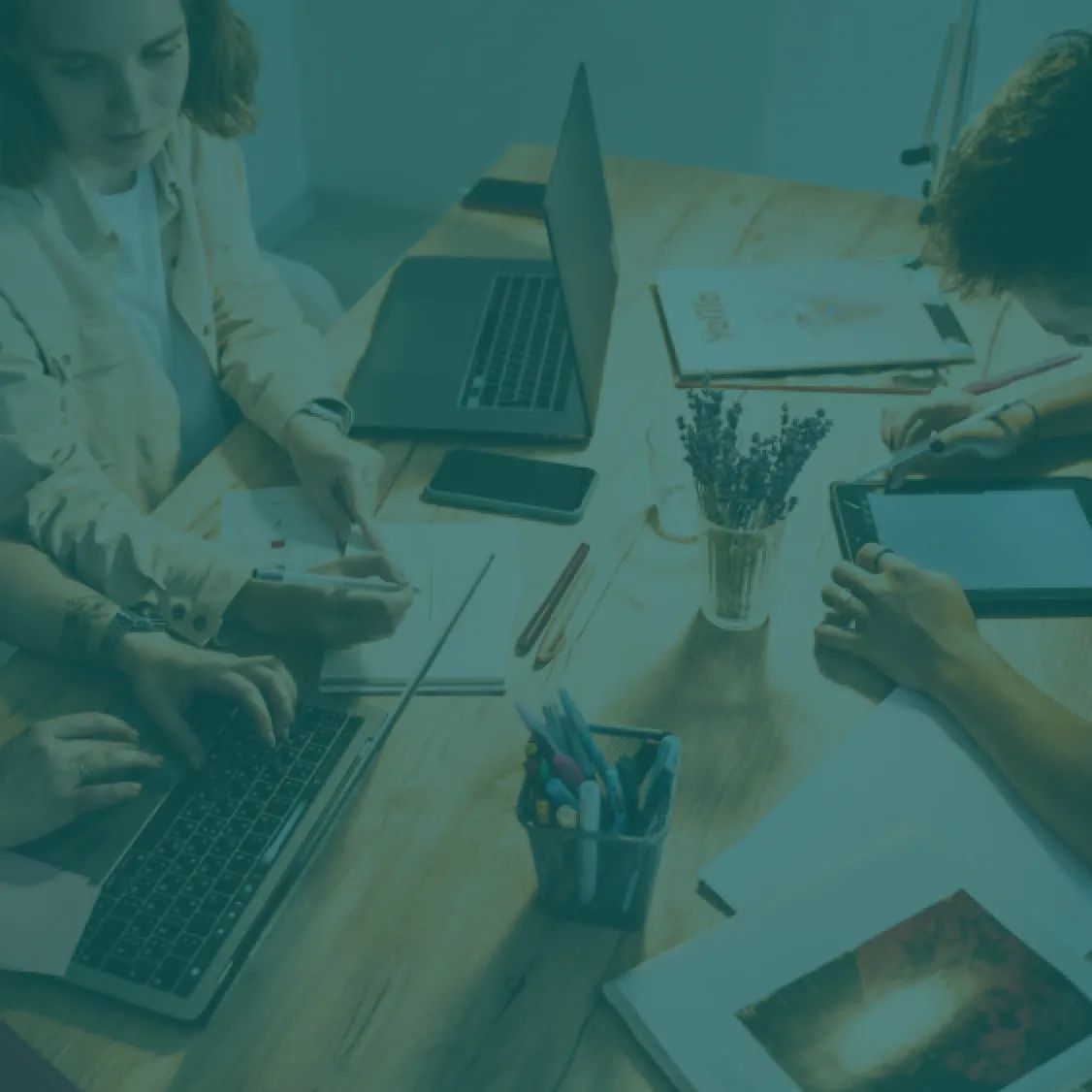 Imagem de fundo em tom verde-petróleo mostrando uma mesa de trabalho com pessoas colaborando, papéis e dispositivos tecnológicos, representando o ambiente de inovação e ética da equipe Fidúcia