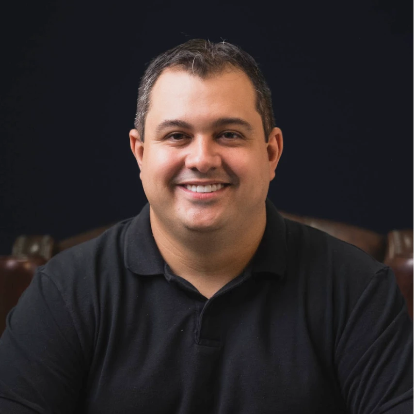 A man reading from a tablet in a collaborative office environment. Desks with documents and a discussion between colleagues are visible in the background.