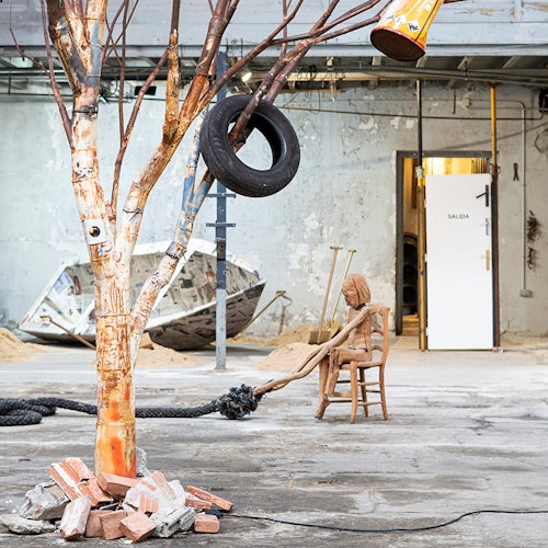 Taller abandonado con un columpio de neumático en un árbol pintado, una figura de madera en una silla, ladrillos esparcidos y una puerta abierta con el letrero "Salida."