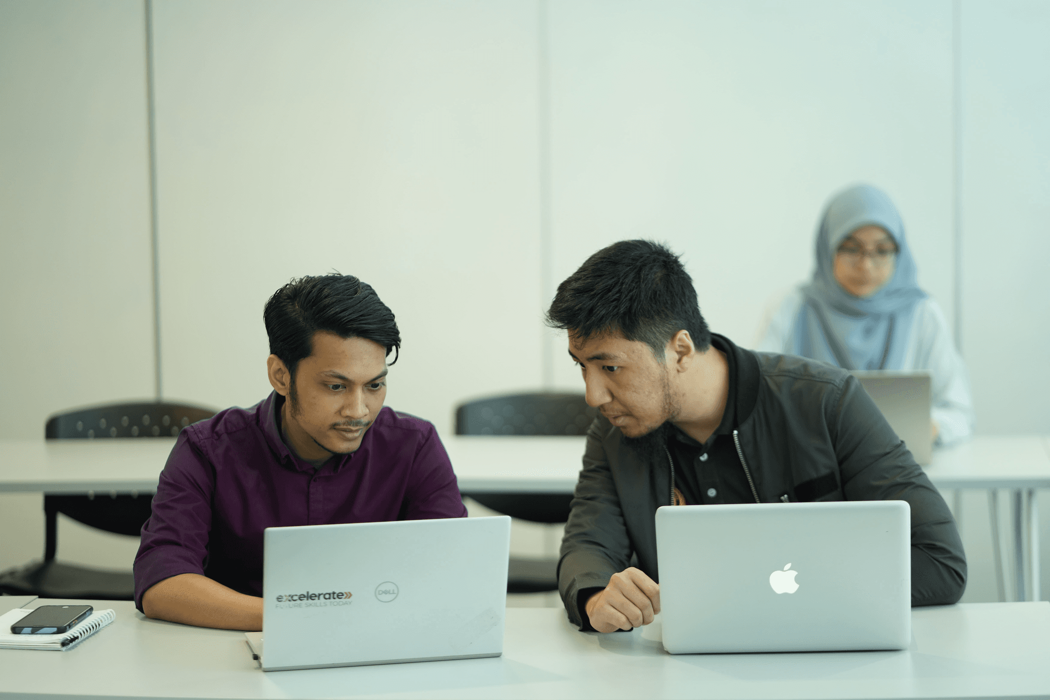 1. Two men seated at a table, each working on their laptops, engaged in a collaborative discussion.