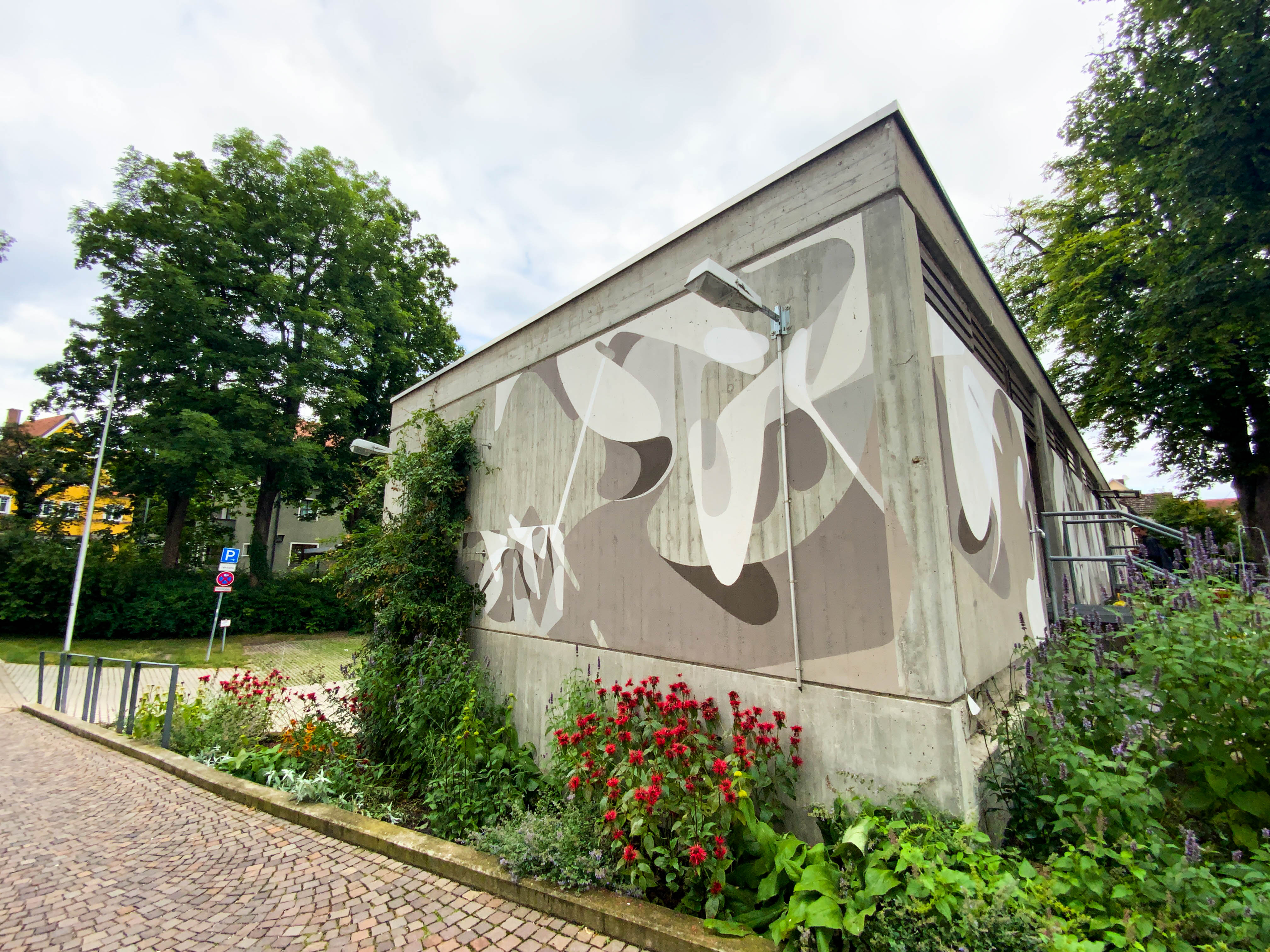 An abstract monochrome mural painted on a cube-shaped building, surrounded by green shrubs and red flowers beside a pathway.