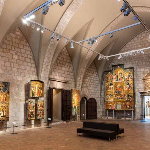 An art gallery with arched ceilings displaying large medieval religious paintings on stone walls with a black bench in the center.