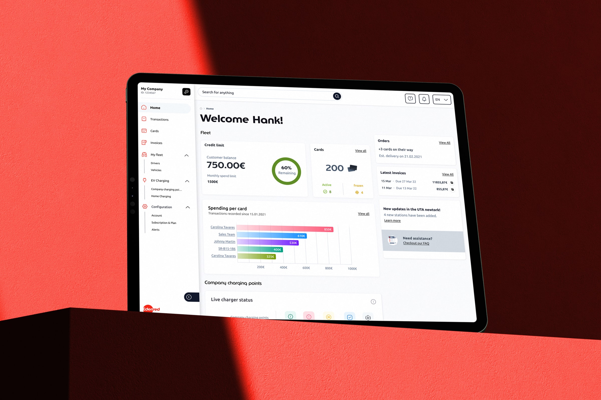 The UTA Edenred fleet management dashboard displayed on a tablet device, photographed at an angle against a dramatic red and dark background. The screen shows the personalised home screen with a welcome message, fleet credit limit, card status, spending per card bar chart, latest invoices, and a live charger status section.