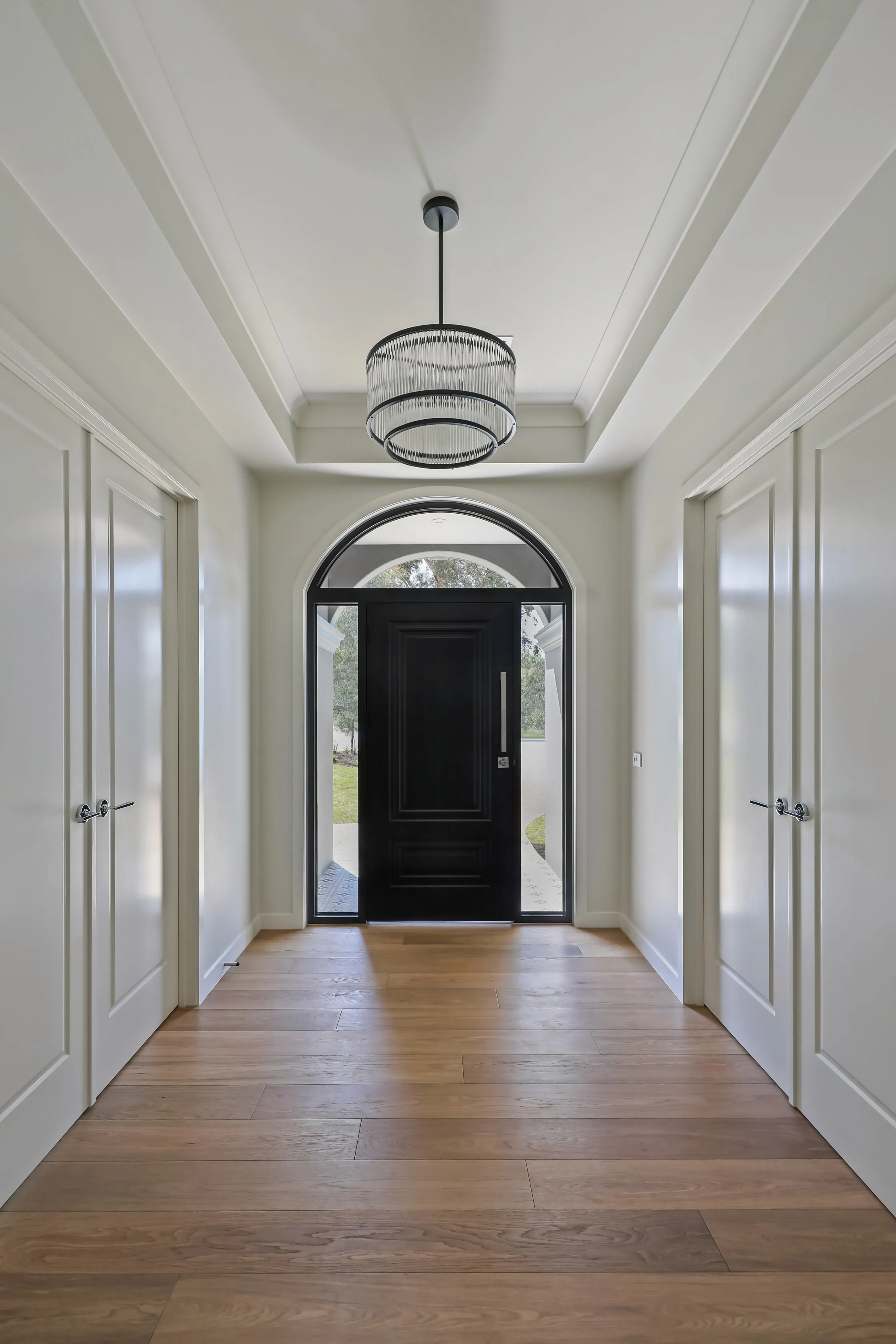 Entry hallway with a tray ceiling and ribbed-glass drum chandelier, white panelled doors, oak floors and an arched black front door with sidelights to the garden