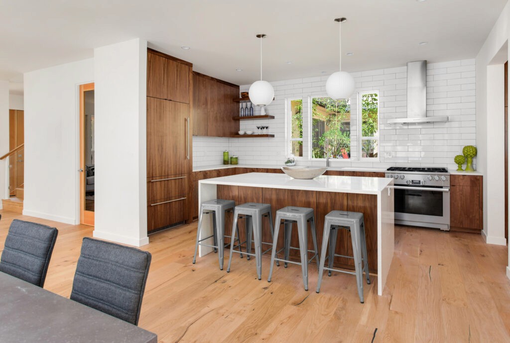 Modern kitchen with white walls and backsplash, natural wood cabinets, a white island with a waterfall countertop, and four metal barstools. Two round pendant lights hang above the island, and a stainless steel oven and hood are installed along the back wall with windows above the countertop.