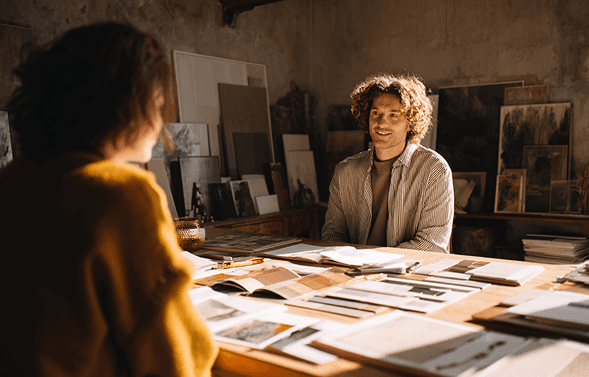 A person sits at a wooden desk in a warmly lit home office, focused on a computer screen, with a lamp, books, and a pencil holder nearby, creating a cozy and productive workspace.