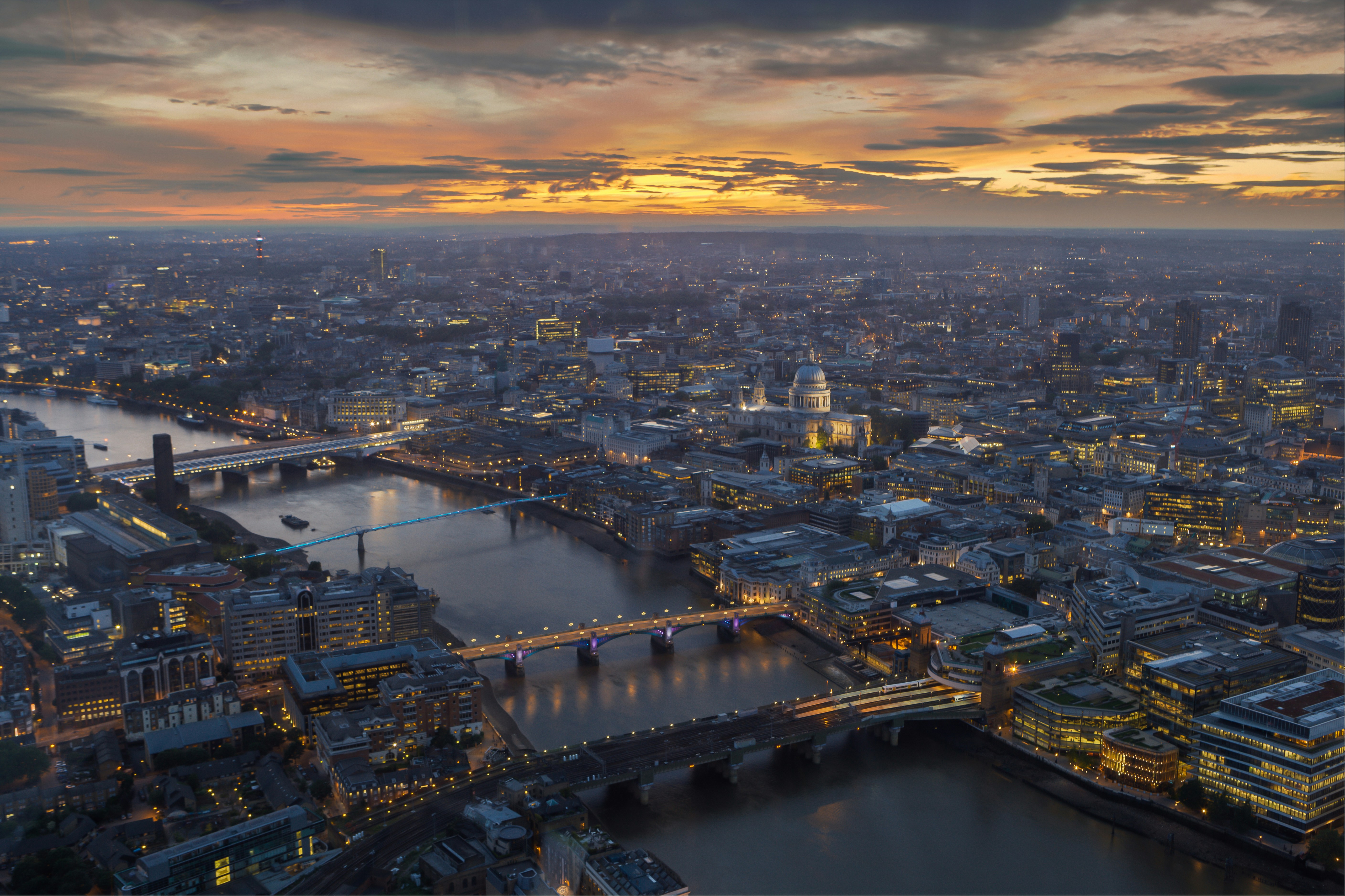 Aerial view of London at sunset, showcasing the illuminated St. Paul’s Cathedral, the River Thames, and several iconic bridges, with the city skyline in the background.