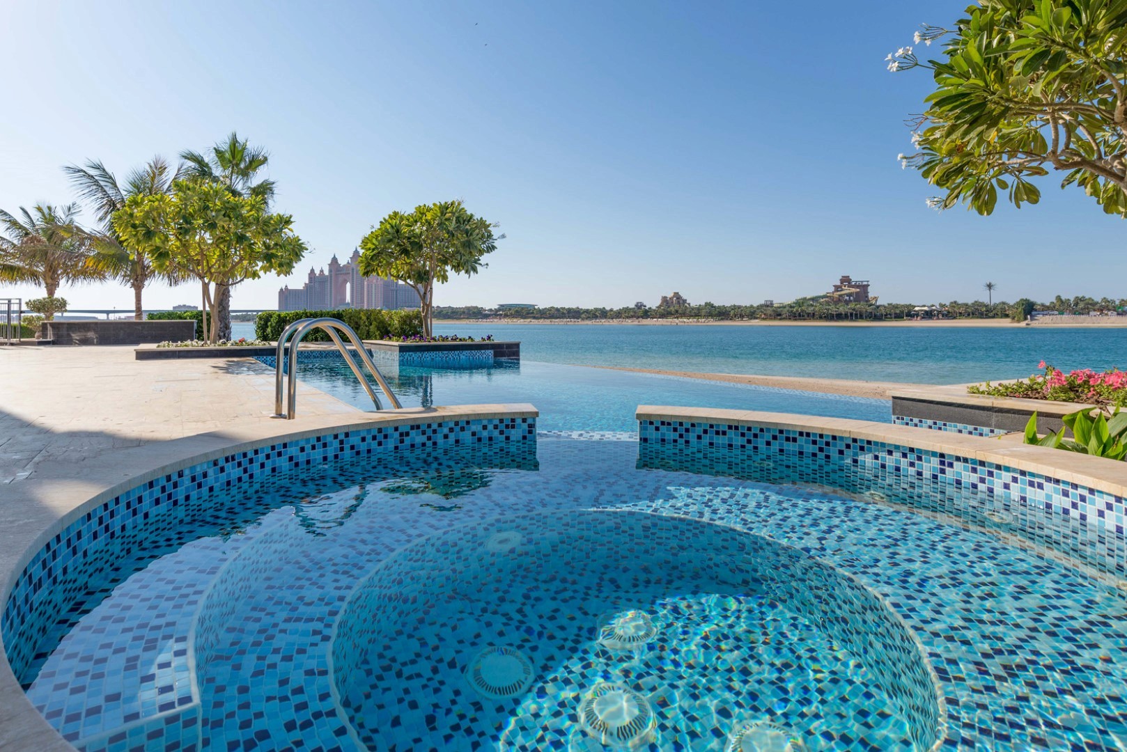 A luxurious infinity pool with intricate blue mosaic tiles overlooks a serene ocean, surrounded by lush palm trees and tropical greenery, with a clear blue sky and distant modern architectural landmarks in the background.