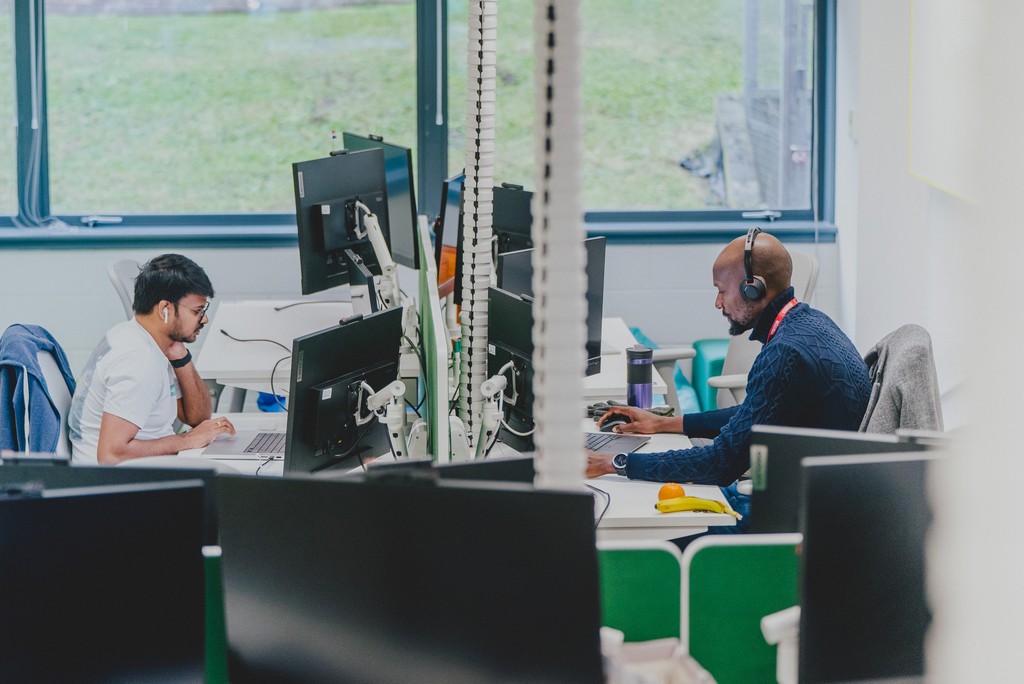 People working at computer desks in an open‑plan office, with screens and desk dividers visible.