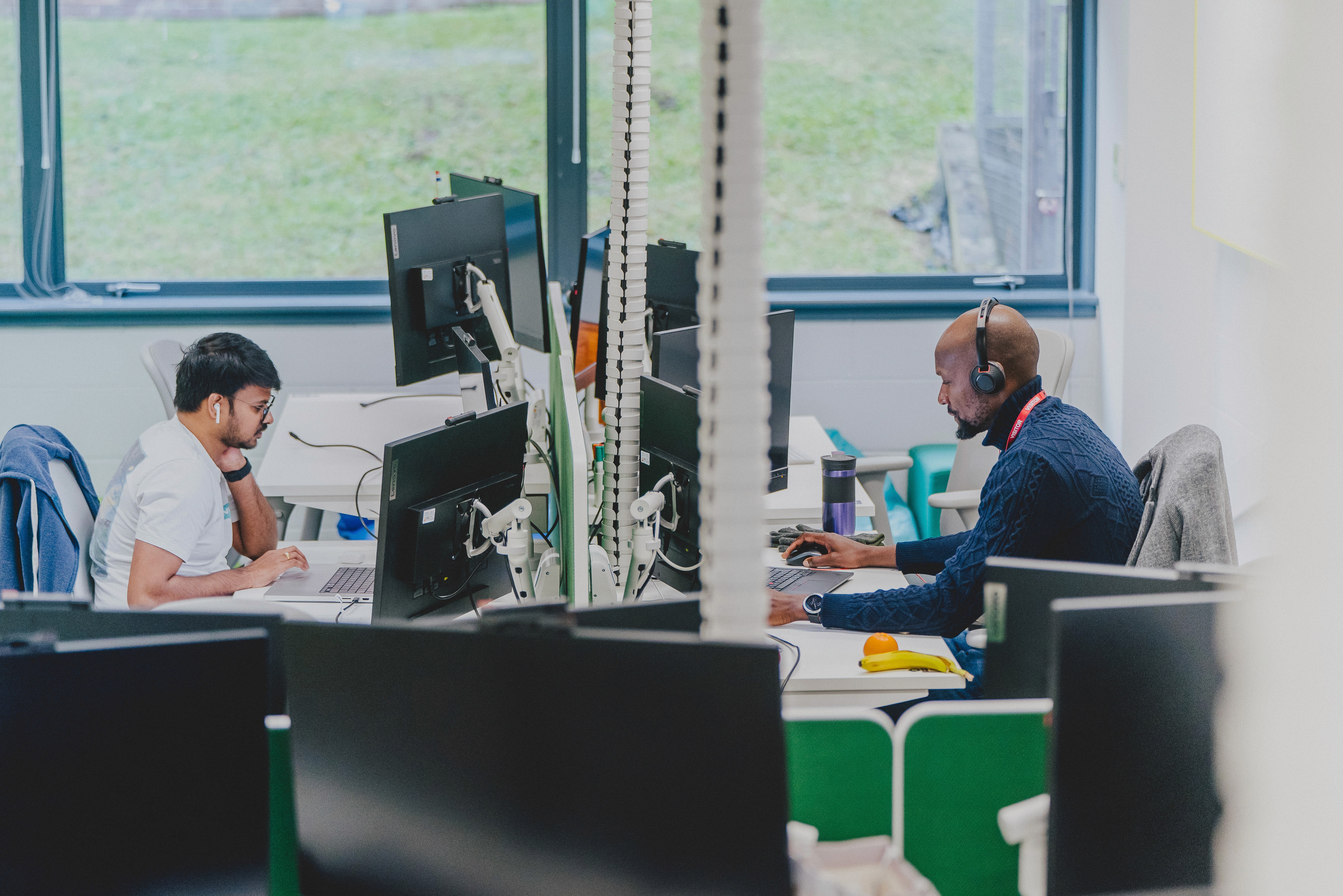 Two people work at adjacent desks in an open-plan office, each using multiple monitors, with windows providing natural light in the background.
