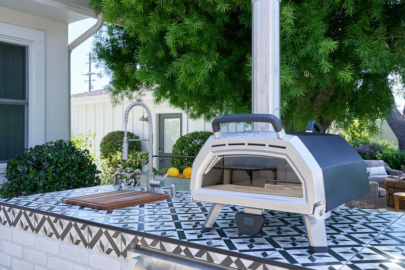 Close-up of an outdoor pizza oven with decorative tile on a countertop, set against a backdrop of lush greenery. Costa Mesa Exterior Remodel by Todd Huge.