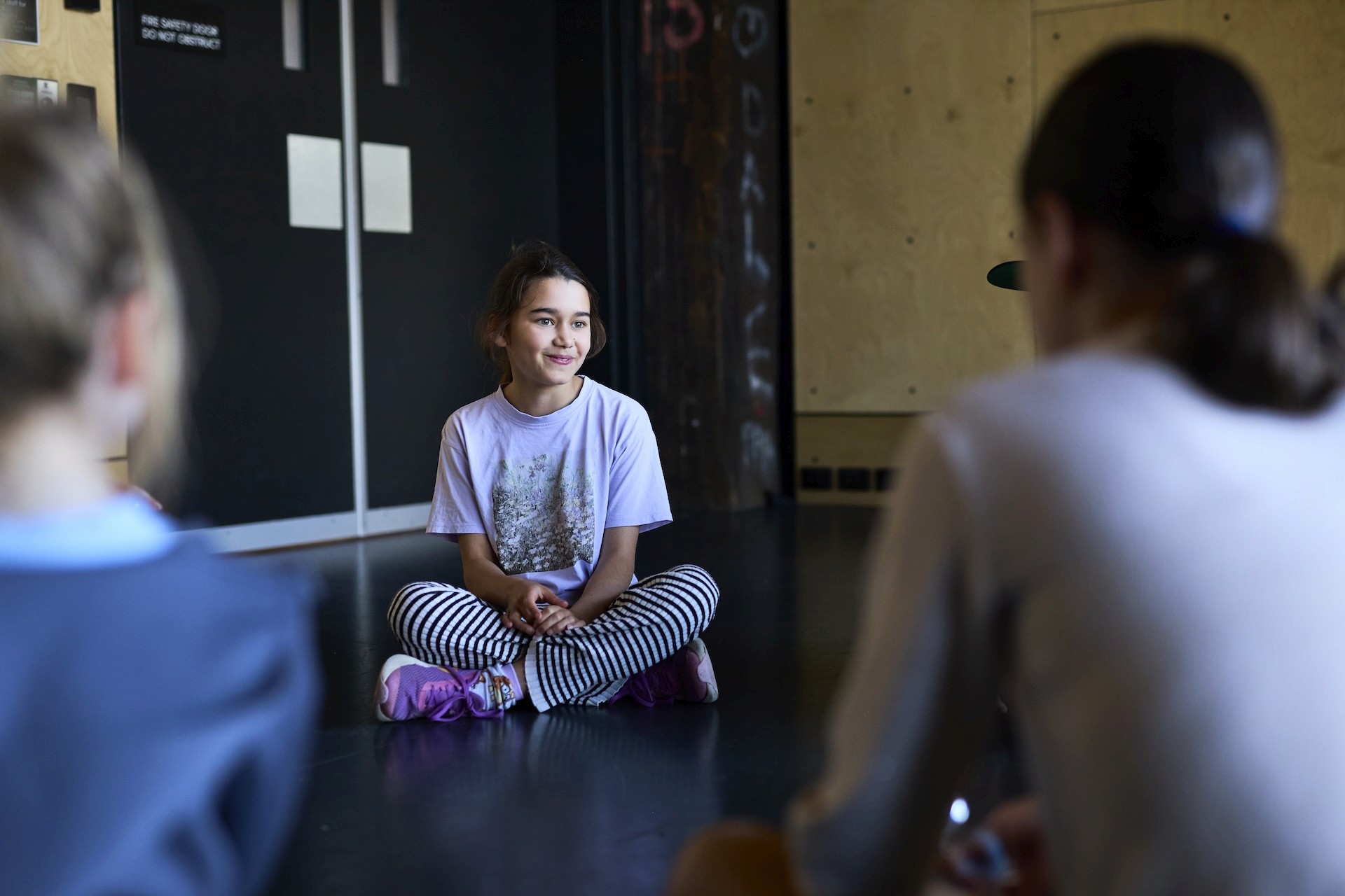A child sits cross-legged on the studio floor, smiling and listening to others during a group activity.