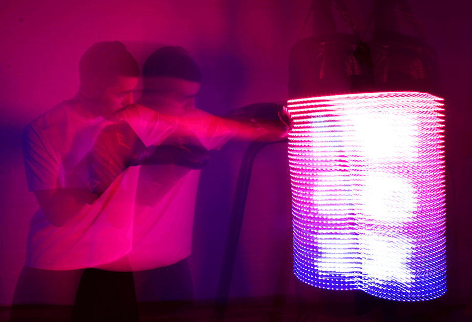 Two individuals are interacting with a large cylindrical light fixture illuminated with pink and blue LED lights. The person on the left, wearing a white t-shirt, reaches toward the fixture with their right hand. The person on the right, wearing a dark shirt, stands slightly behind and to the left of the person on the left, partially obscured by the light. The background is a dark wall, and the lighting casts a pinkish-purple hue across the scene.