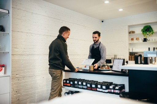 Two people converse in a modern kitchen, discussing something on a counter. Natural light fills the space.