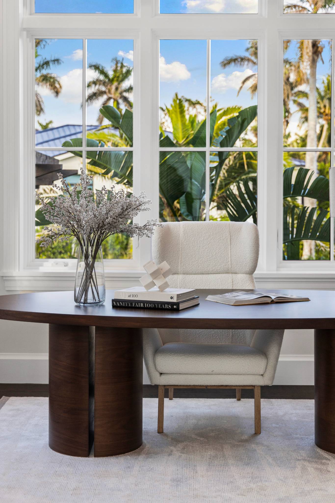 a spruce desk with books laying on top of it and windows with palm trees in the background