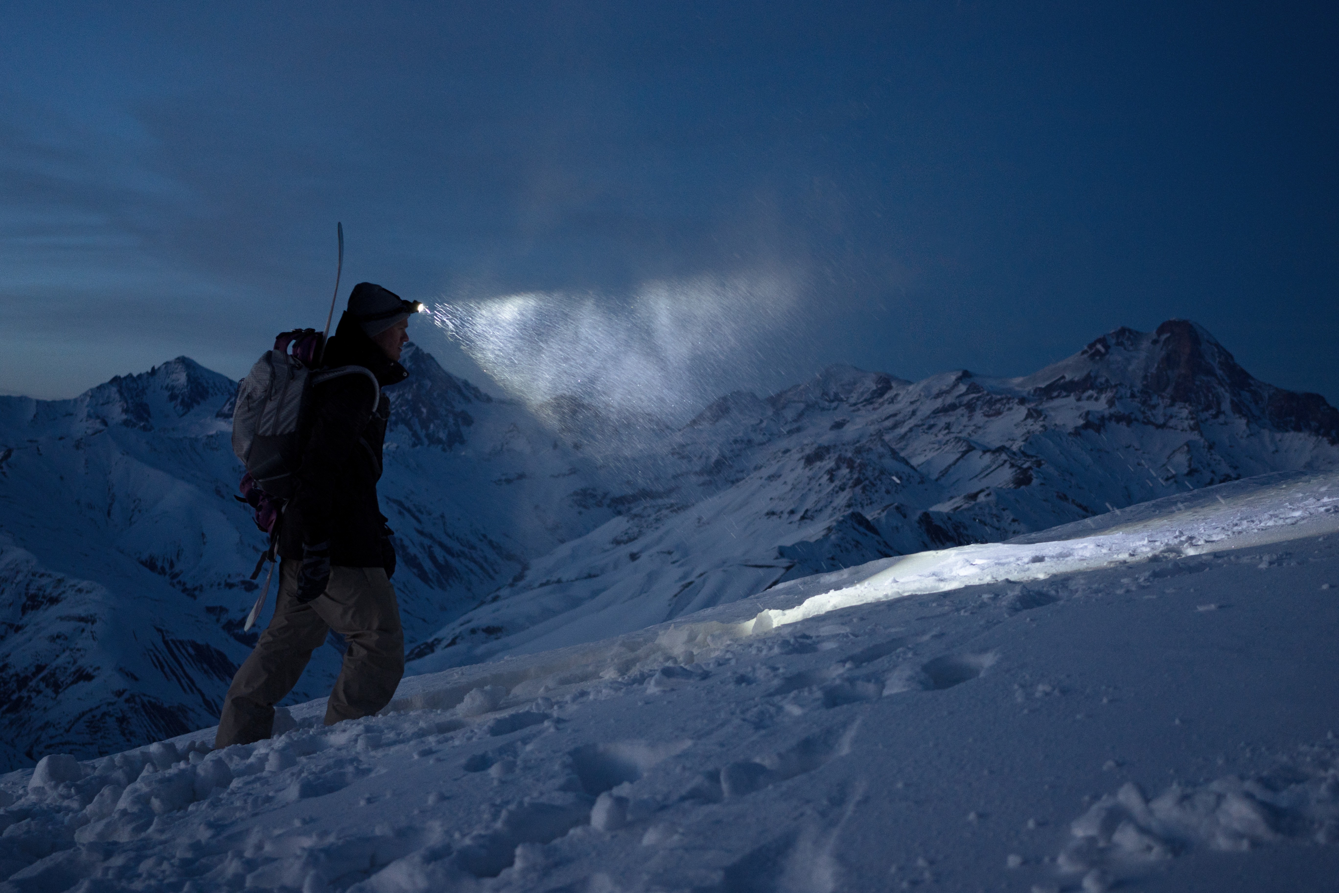 A person hiking uphill on a snowy mountainside. Skis are strapped to the person's back. It's dark and snow is falling, visible in the light of the headlamp.