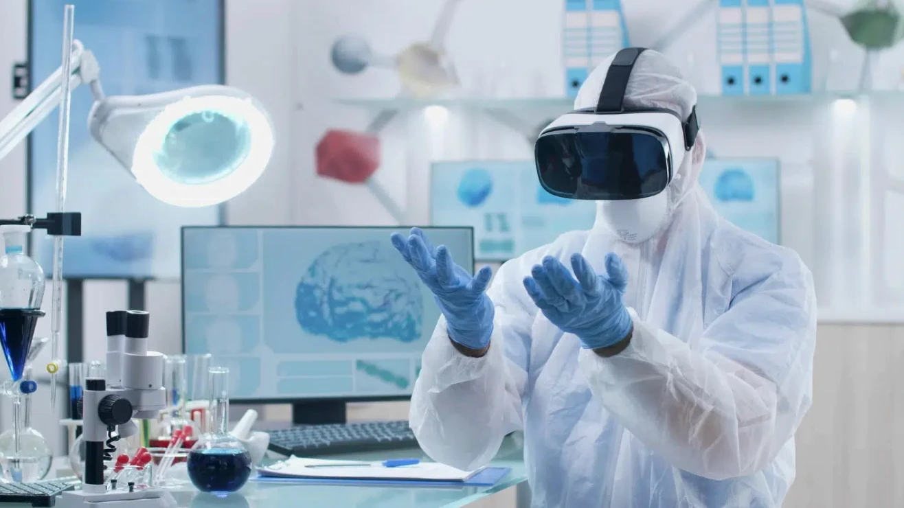 Pessoa vestindo traje de proteção e óculos de realidade virtual em laboratório, com equipamentos científicos sobre a bancada e monitor ao fundo.