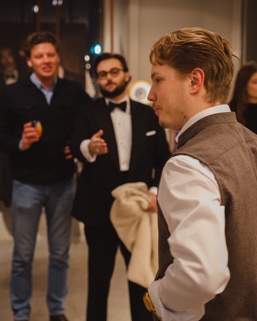Three young men at a social gathering, two dressed in tuxedos and one in a vest and white shirt, engaged in conversation.