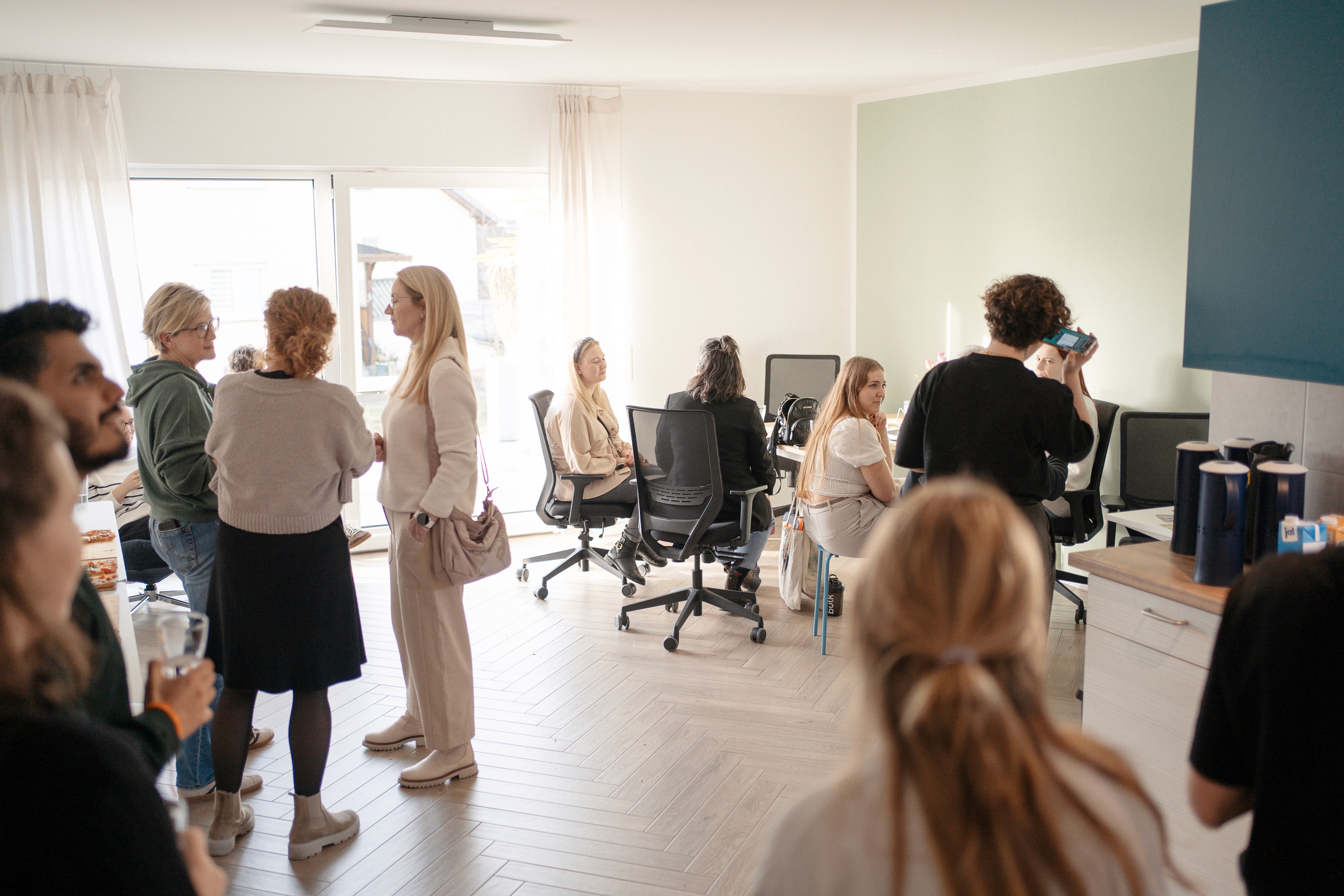 Mehrere Personen unterhalten sich und arbeiten in einem hellen Coworking-Space mit Schreibtischen, Bürostühlen und Küchenzeile