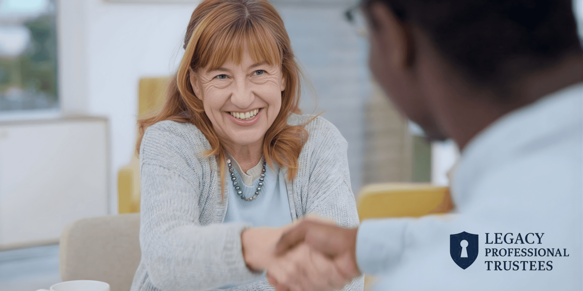 Smiling woman in a cozy office setting shakes hands with a man, conveying collaboration. Logo reading "Legacy Professional Trustees" is visible.