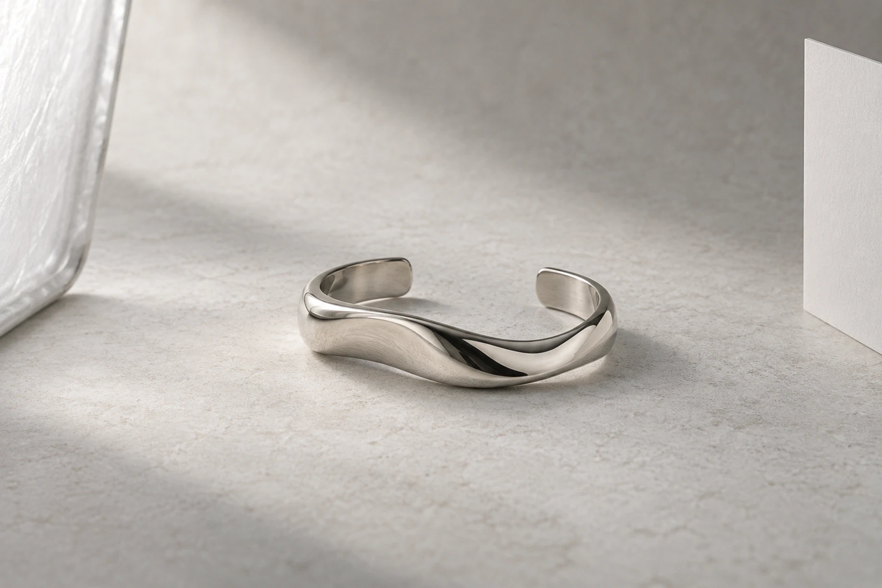 Polished silver cuff on a pale studio surface between a diffuser edge and a white card