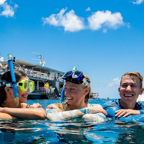 三個戴著潛水裝備的人在陽光明媚的藍天下，在一艘船附近的海洋中微笑和休息。