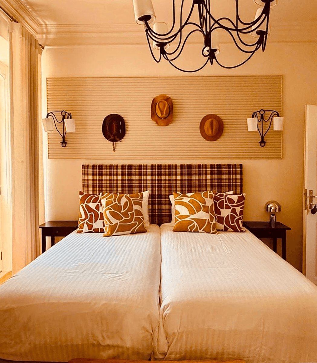 Cozy bedroom with a checkered red-and-white headboard, two pillows, a brown hat on a blanket, a bedside lamp, and an open door, exuding warmth.