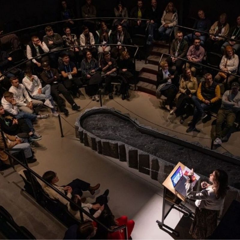 A speaker stands at a podium addressing a full audience seated in a circular, tiered showroom at Lava Show, presenting during an Innovation Week event in Reykjavík.