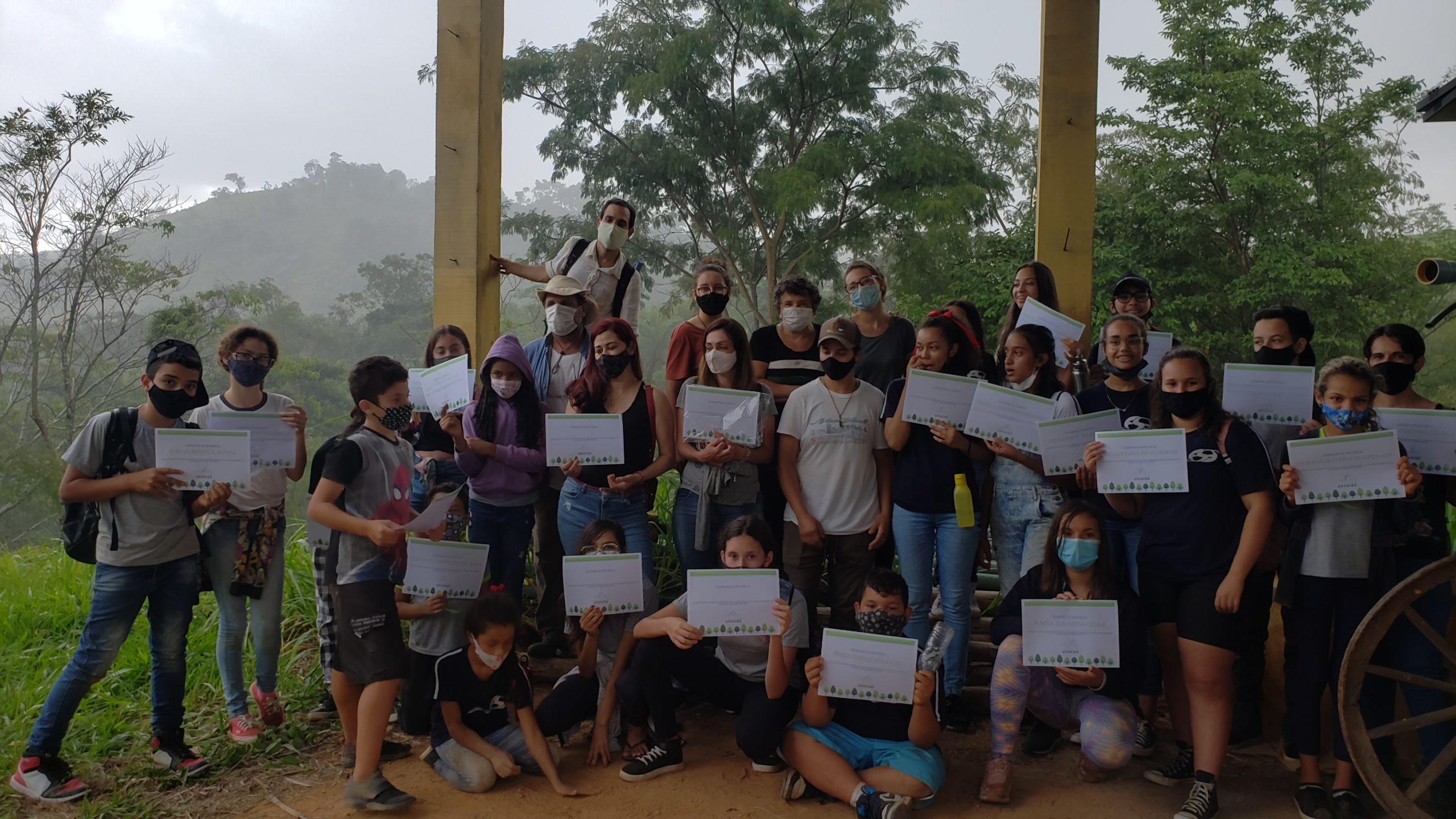 Grupo de alunos de máscara segura folhas impressas em área coberta cercada de mata e morros