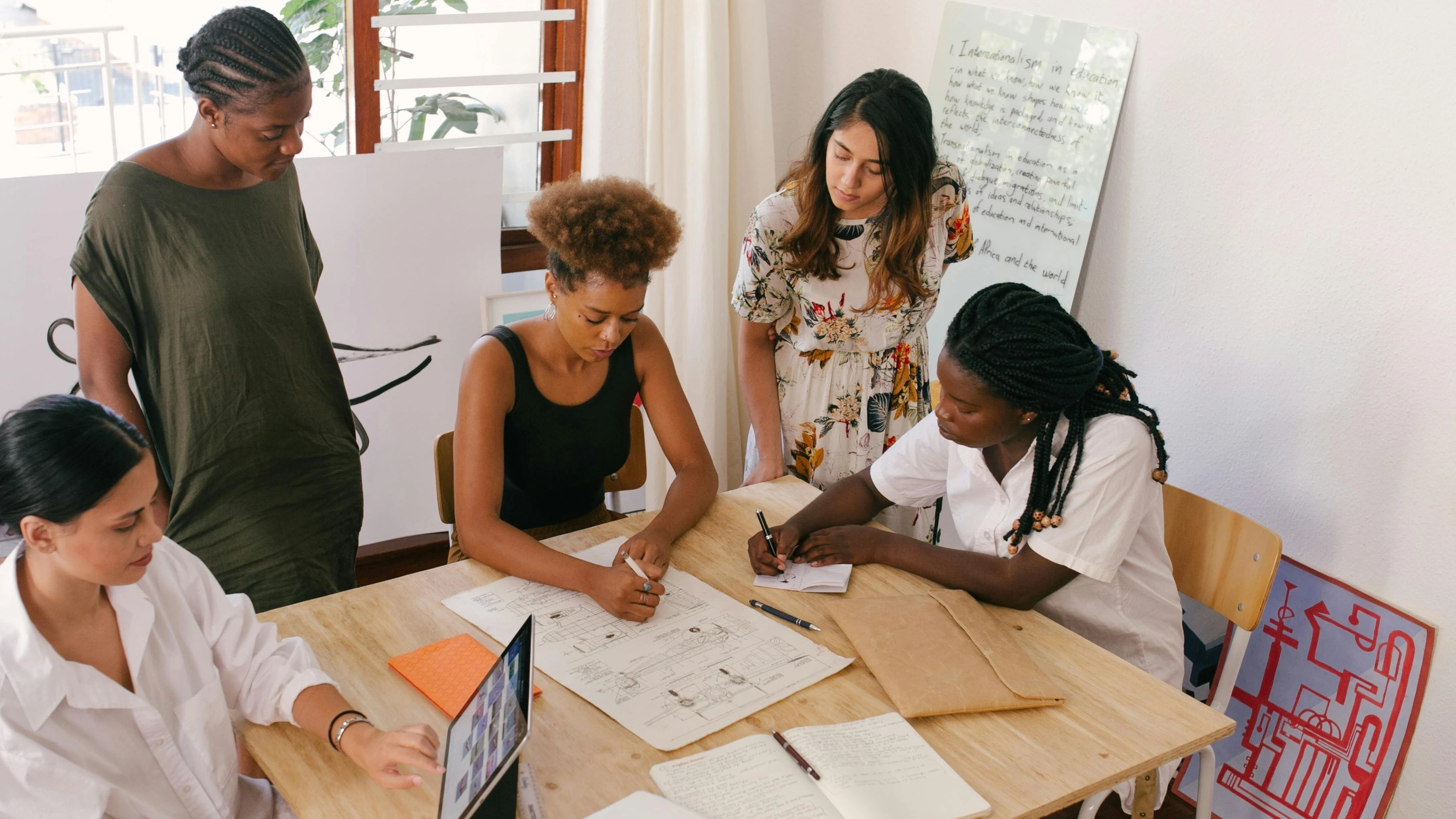 Diverse group of young women collaborating on a creative project, planning and designing with sketches, laptops, and whiteboards in a modern workspace.