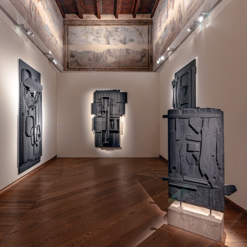 Una galleria presenta tre sculture astratte dai toni scuri montate su pareti di colore chiaro, con un soffitto in legno e faretti.