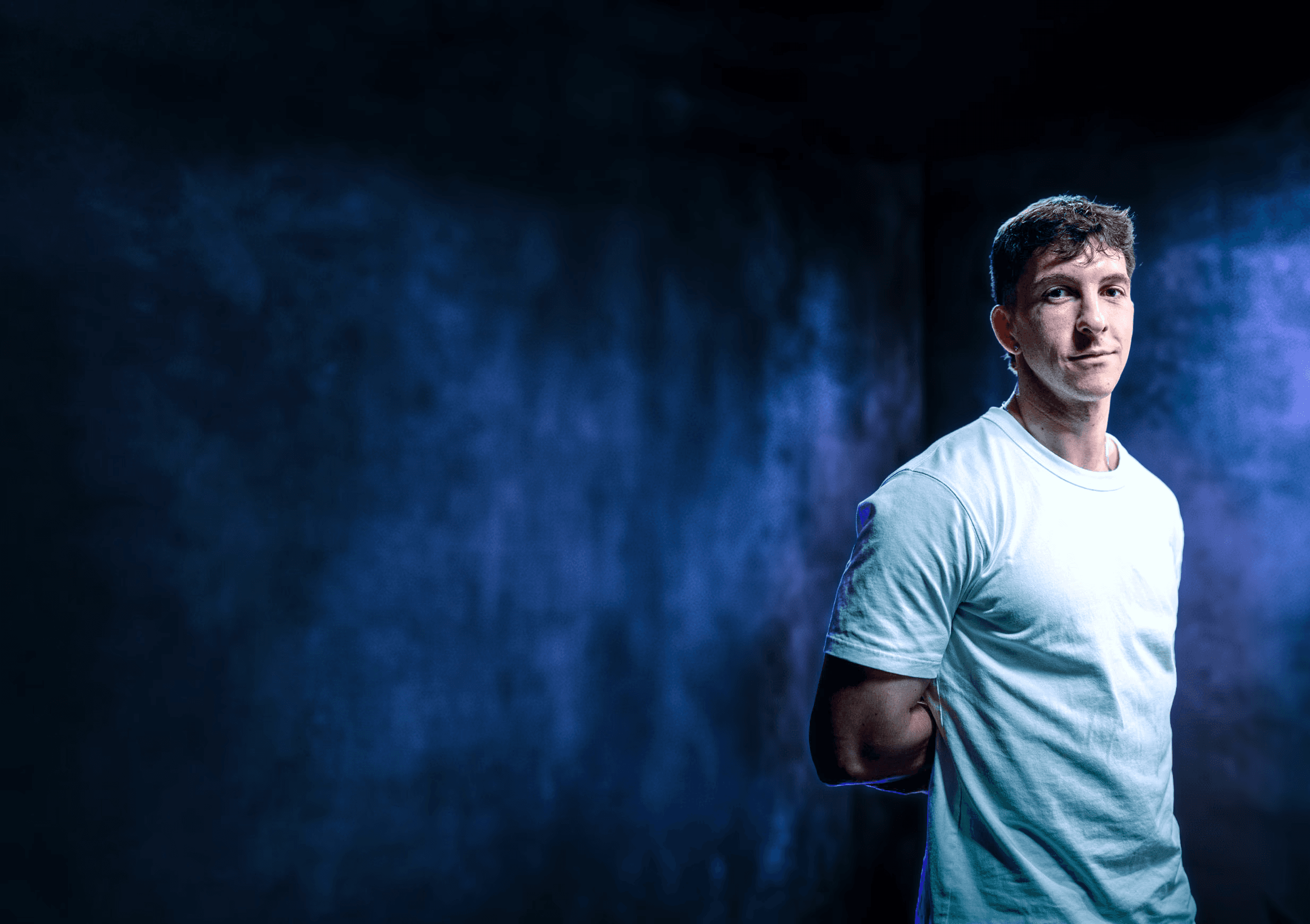 Portrait professionnel et moderne d'un jeune homme, représentant le fondateur ou directeur artistique de Nux-Studio, posant de face avec un léger sourire. Il porte un simple t-shirt blanc à manches courtes. L'arrière-plan est un mur texturé, sombre et baigné d'un éclairage dramatique de couleur bleu électrique et violet, créant une ambiance de studio créatif et innovante. L'espace négatif important sur la gauche est idéal pour l'ajout d'un titre, d'un logo, ou d'un slogan. Ce visuel évoque l'expertise en branding, direction artistique et l'identité moderne d'une agence de design digital.