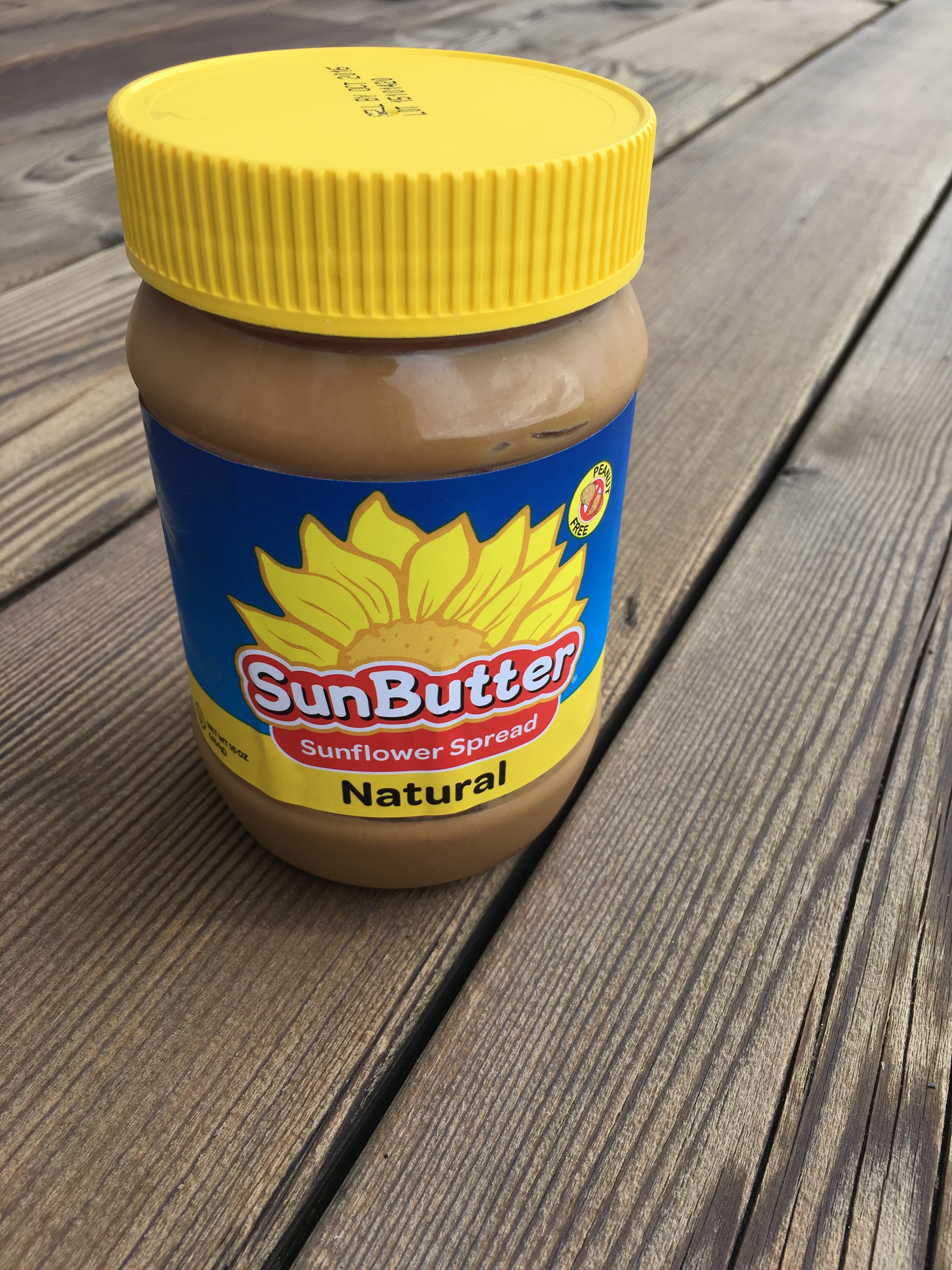A jar of SunButter Natural sunflower spread with a yellow lid and vibrant blue label featuring a sunflower logo is placed on rustic wooden planks.