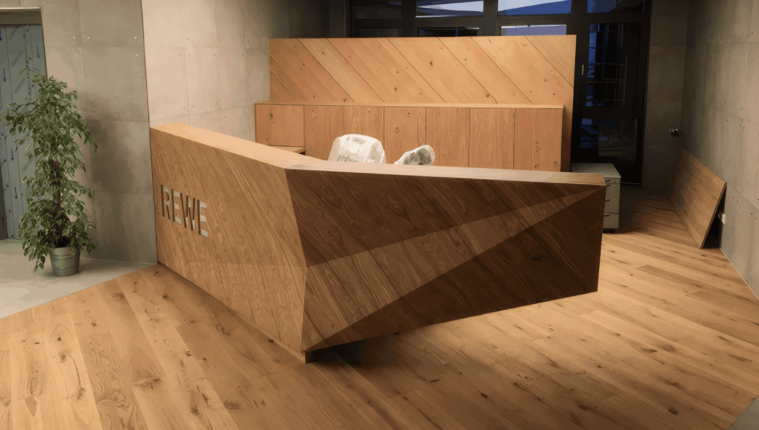 MW Interior - REWE Empfangstresen aus Massivholz mit diagonalen Holzpaneelen im Foyer" Beschreibung: Eindrucksvoller Empfangstresen in geometrischer Holzoptik mit eingelassertem REWE-Schriftzug – ein skulpturales Designelement im Eingangsbereich auf Eichenparkett-Boden.