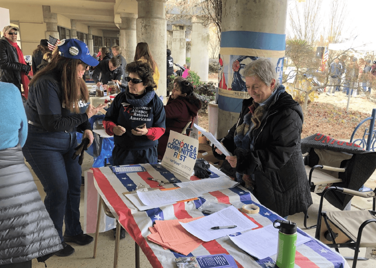 Three women engage at an information booth covered with papers and a patriotic tablecloth, promoting “Schools & Communities First.”