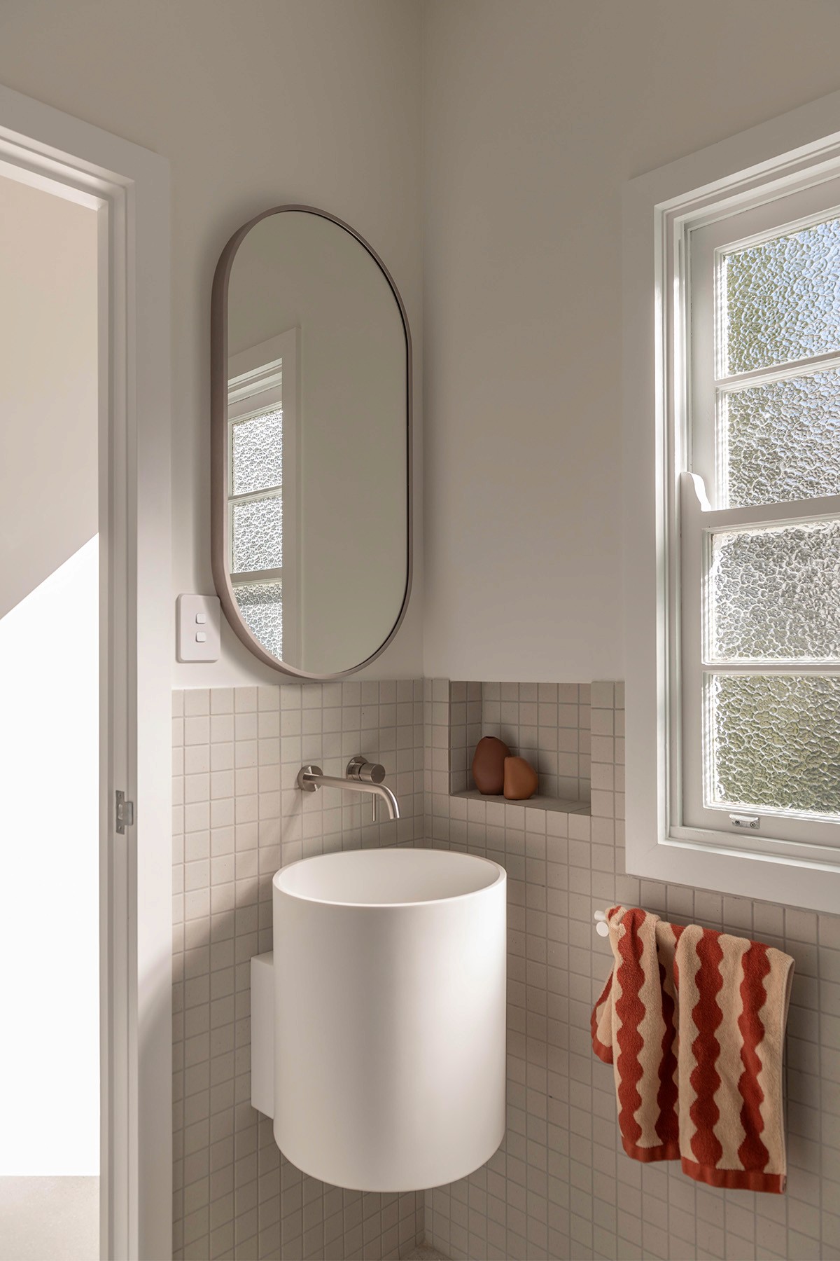 Compact bathroom basin with wall-mounted cylindrical vanity, oval mirror, and frosted window filtering soft daylight.
